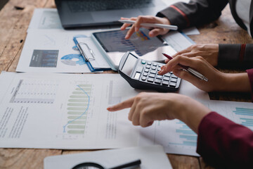 Two businesswomen are working together, analyzing financial charts, using a calculator, and discussing data on a tablet in a collaborative office meeting