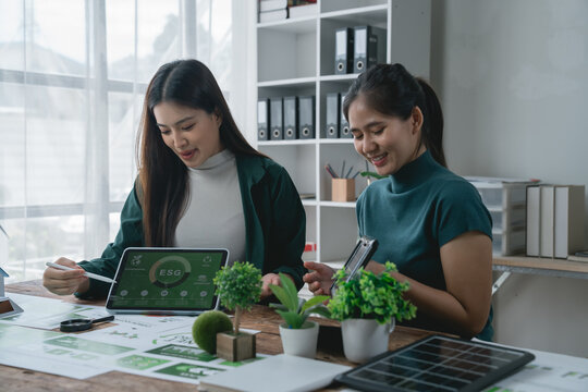 Two Asian businesswomen smiling and collaborating on an ESG project, analyzing a solar panel and using a tablet with a graph in a modern office filled with plants - Powered by Adobe
