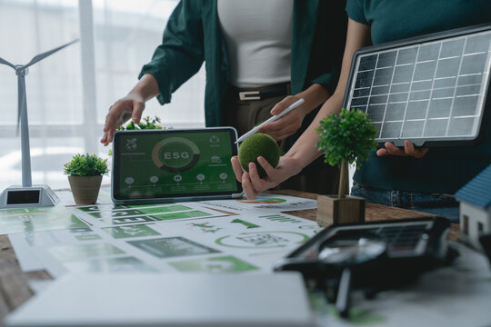 Businesswomen holding a solar panel and a moss ball discussing an ESG project with charts, a tablet displaying ESG infographic, and a wind turbine model on a wooden table - Powered by Adobe