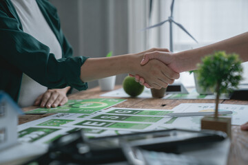 Two businesswomen shaking hands after successfully closing a deal on an ESG project, surrounded by documents, a miniature wind turbine, and a tree on a wooden table