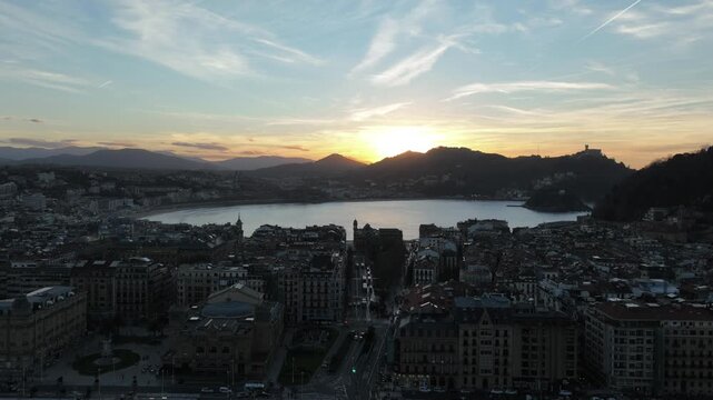 Drone flight over the city of San Sebasti&aacute;n towards the iconic La Concha Beach, capturing a stunning golden-hour sunset over the bay and the rooftops of the Basque coast in northern Spain