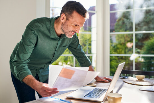 Smiling man doing paperwork at home using laptop