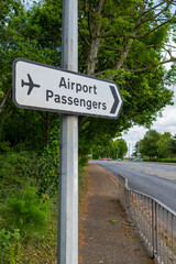 White directional road sign with aeroplane icon pointing to the Airport Passengers area. Green trees and a rural road create a peaceful transportation wayfinding scene