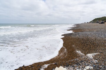 Pebble beach with white foam waves washing ashore at Cromer, Norfolk, England. Dramatic coastal landscape with shingle shoreline and cloudy sky over the North Sea