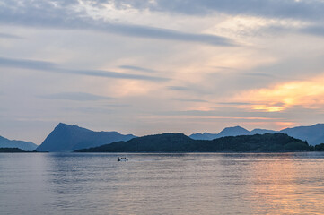 As the sun sets, a small boat glides across the calm sea, surrounded by mountains under a pastel sky.