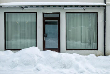Snow blankets the ground in front of a vacant storefront with large windows and a wooden door, creating a quiet winter atmosphere.