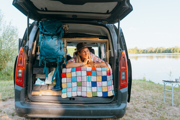 A solo blonde woman wearing a hat sits comfortably in her mini campervan enjoying vanlife freedom and calm