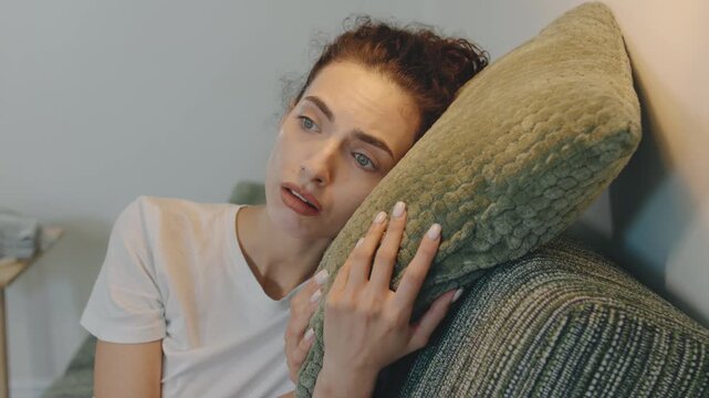 A person sits on a couch in a warmly lit room, looking thoughtful and sad with her head resting on a green pillow, reflecting on her emotions.
