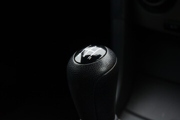 Close-up shot of a manual car gearstick, showing the gears and reverse. The focus is sharp on the black leather-textured knob with white markings.