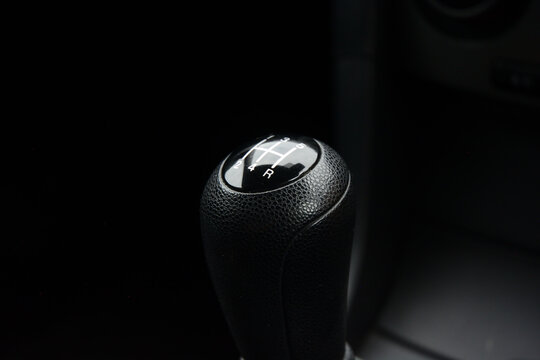 Close-up shot of a manual car gearstick, showing the gears and reverse. The focus is sharp on the black leather-textured knob with white markings.