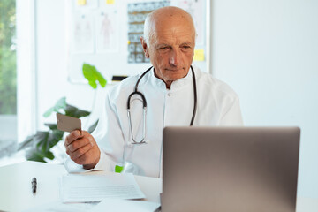 Old male doctor in medical uniform holding credit card making shopping online at clinic