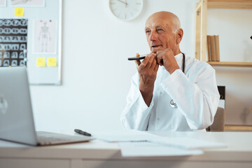 Elderly senior doctor in uniform recording voice message using smartphone in hospital office