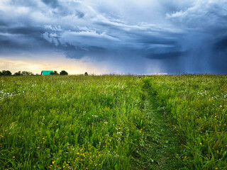 Stormy clouds gather over a lush green field with a distant house at twilight