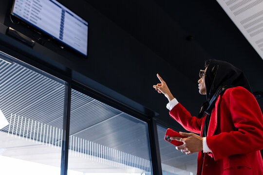 Businesswoman pointing at airport information board while holding smartphone