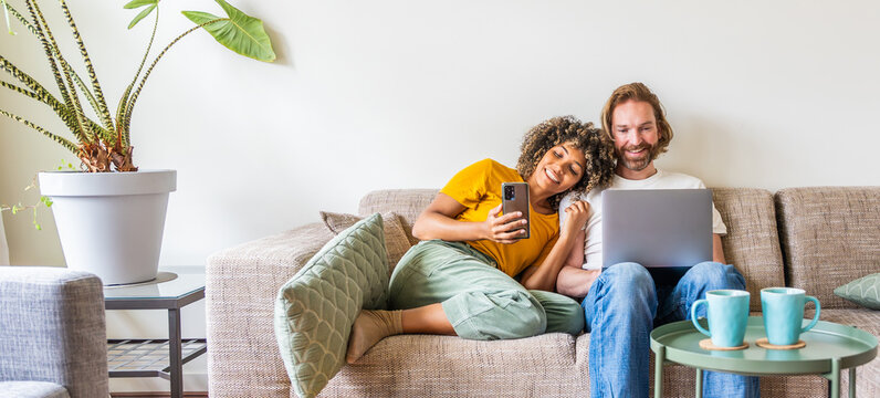 Multiracial young couple using computer laptop and smart mobile phone sitting on the sofa at home - Happy diverse husband and wife using pc online services - Technology life style concept