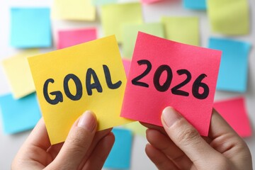 A person holds up a bright yellow sticky note with the word goal and the year 2026 written in bold black letters, surrounded by colorful notes in a festive New Year atmosphere