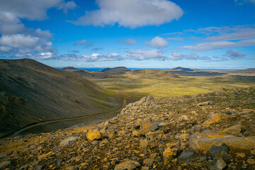Vast Icelandic highlands open toward the horizon, where rugged volcanic slopes meet golden plains under a wide blue sky filled with drifting clouds
