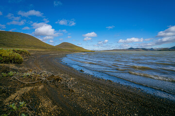 Volcanic black sand shoreline of a highland lake with rolling hills and blue skies in Iceland