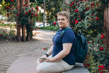 Bottom view smiling happy fun young man in blue t-shirt sit on bench use mobile cell phone chat online relax in green city sunshine park outdoors. Urban lifestyle leisure concept.