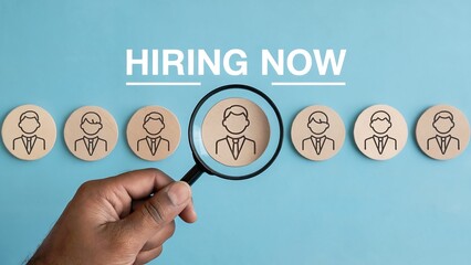 Hand holding a magnifying glass over a row of employee icons, with hiring now text above, symbolizing recruitment and talent search