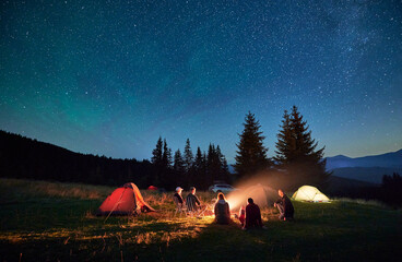Group of friends hikers having a rest around campfire, surrounded by glowing tents under...