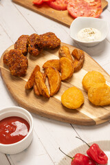 Fried potatos and nuggets served on a wooden board on a white wooden table. More ingredients surround them: ketchup and mayonnaise, sliced tomatoes and chilli pepper. 45 degree view