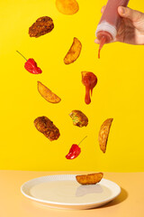 Potato wedges, nuggets, ketchup and red spicy peppers over a white dish with a yellow background and a hand squeezing a bottle of ketchup.