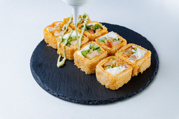 Portion of deep-fried sushi rolls with vegetables and cream cheese, drizzled with sauce, served on a black stone plate.