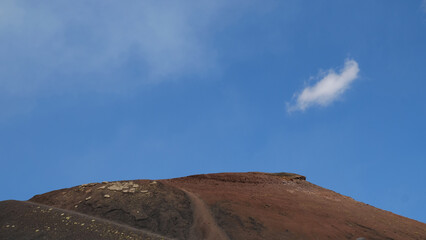 Striking view of a volcanic crater on Mount Etna. Reddish-brown soil against bright blue sky. Faint path leads up to the peak, symbolizing journey or challenge. Travel, nature, geology.