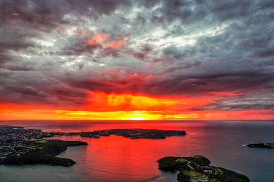 Aerial view of the fiery sunset reflecting on the tranquil waters near Balmoral Beach, where the sky meets the land in a dance of light and shadow, Balmoral Beach, New South Wales, Australia.