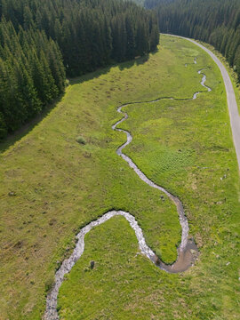 Aerial view of a winding stream cutting through a vibrant green meadow, bordered by a dense forest and a road, Sihla, Bansk&Atilde;&iexcl; Bystrica Region, Slovakia.