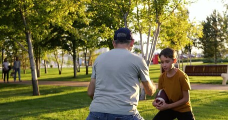 Happy father and son playing rugby on green park lawn at sunset. Dad teaches his boy skills while son enjoys game. Father and child playing rugby brings joy, teamwork and family bonding. Warm light. - Powered by Adobe