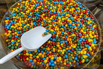 Colorful candy-coated chocolate in bulk display. Large container filled with assorted colorful candy-coated chocolates and a white scoop, typically found in candy shops or markets.