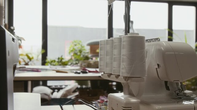 Medium no people shot of professional industrial sewing serger machine on worktop in atelier, with large bobbins of white thread, filmed against cutting table and large window at daytime