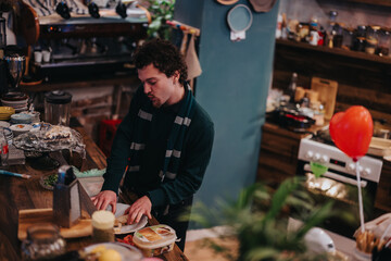 A man is cutting bread and preparing food in a comfortable, rustic kitchen with warm lighting.