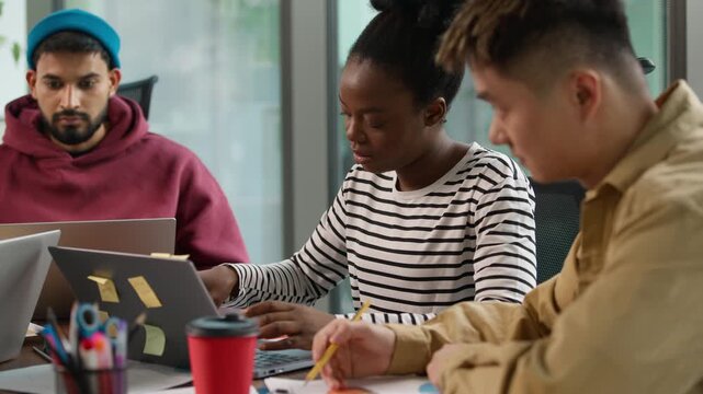 Three multiracial casual informal business employers at table indoors in corporate office looking in documents review data diagram laptop use startup development team busy concentrated workgroup work
