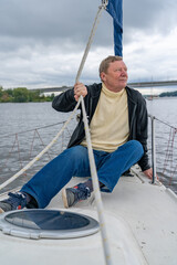 Mature man in yellow turtleneck and black hoodie relaxing on bow of sailboat, holding rope, and gazing thoughtfully at serene water and distant bridge under overcast sky.