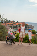 Family vacation photo with mother and her two kids, one in stroller, overlooking beautiful Mediterranean coastal town with red roofs and clear blue water.