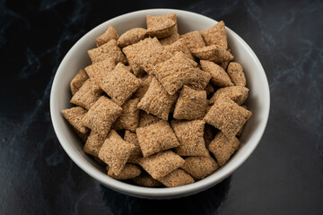 A white bowl filled with crunchy whole grain cereal squares sits on a dark surface, perfect for a nutritious breakfast or a quick snack option.