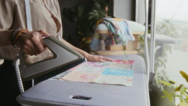 Medium close-up of hands of young seamstress picking up hot iron and pressing button to test steam, before pressing patchwork fabric piece and smoothing out creases after stitching
