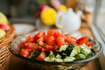 Glass bowl containing freshly chopped tomatoes and cucumbers resting on wooden kitchen table near woven apple basket and vintage teapot, representing light, nutritious meal preparation