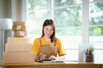 A young Asian female entrepreneur works from home, managing online sales, shipping parcels, marketing products, and ensuring client satisfaction efficiently.