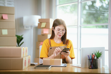 A young Asian female entrepreneur works from home, managing online sales, shipping parcels, marketing products, and ensuring client satisfaction efficiently.