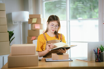 A young Asian female entrepreneur works from home, managing online sales, shipping parcels, marketing products, and ensuring client satisfaction efficiently.