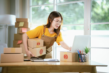 A young Asian female entrepreneur works from home, managing online sales, shipping parcels, marketing products, and ensuring client satisfaction efficiently.