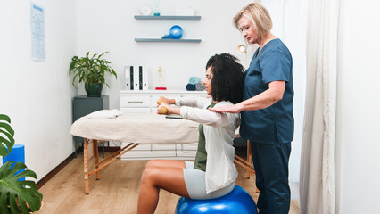 A Senior Physiotherapist Assists a Young Woman Client with Strength Training Exercises Using Dumbbells and an Exercise Ball in Physical Therapy Clinic