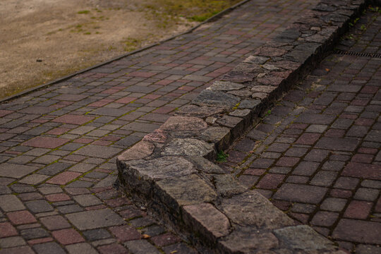 A close-up view of a pedestrian pathway featuring interlocking red and gray bricks and a low stone curb. Moss grows between the gaps, adding a natural texture to the well-worn urban surface.