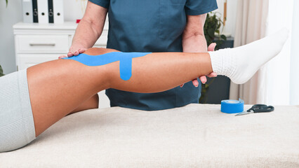A Senior Physiotherapist Skillfully Applies Kinesiology Tape to a Young Black Client's Knee...