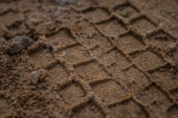 Detailed macro view of a vehicle tire tread imprint in sandy dirt, showing texture and geometric patterns left on the ground.