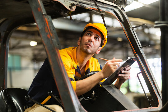 Industrial engineer in safety gear inspecting note clipboard in hand sitting on forklift, working inside factory or warehouse. Focus professional assessing equipment for safety maintenance compliance.
